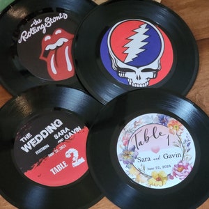May include: Four black vinyl record coasters with different designs. The first coaster features the Rolling Stones logo, the second features the Grateful Dead logo, the third features a wedding announcement with the text "The Wedding Featuring Sara and Gavin June 22, 2024 Table 2", and the fourth features a floral design with the text "Table 1 Sara and Gavin June 22, 2024".
