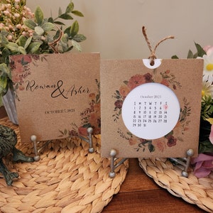 May include: A brown card with a floral design and the text "Rowan & Asher" and "October 7, 2023". A second brown card with a floral design and a calendar for October 2023. Both cards are on a stand.