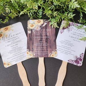 May include: Three wedding fan programs with floral designs. The fans are made of paper and have wooden handles. The text on the fans includes the names Carly Jackson and Carson Maxwell, the wedding date, and the ceremony and reception schedule.