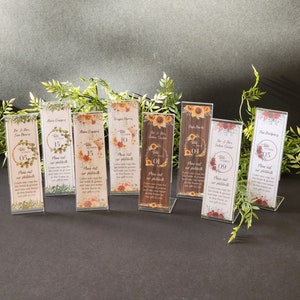 May include: A set of ten clear acrylic table number holders with floral designs and printed text. The table numbers are 01, 04, 05, 09, 10, 15, and 15. The text on the holders includes the names of the wedding couple and the words "Please visit our photobooth".