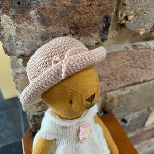 May include: A handmade doll wearing a white crocheted dress with a pink flower accent and a light brown crocheted hat.