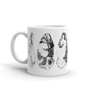 Può includere: Una tazza in ceramica bianca con un'illustrazione in bianco e nero di quattro strumenti in pietra realizzati a mano. Gli strumenti hanno la forma di punte di freccia e sono disposti in fila.