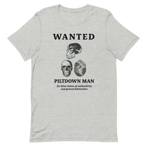 May include: Light gray t-shirt with a graphic design featuring the word "WANTED" above three skull illustrations. Below the skulls, the text reads "PILTDOWN MAN" and a description of the subject's alleged crimes.