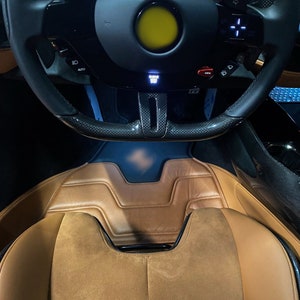 Può includere: Una vista ravvicinata dell'interno di un'auto di lusso. Il volante è in pelle nera con dettagli in fibra di carbonio e presenta un pulsante giallo. I sedili dell'auto sono in pelle marrone con una texture simile al camoscio.