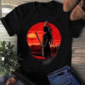 Może przedstawiać: Czarny t-shirt z krótkim rękawem z nadrukiem graficznym przedstawiającym sylwetkę osoby w pióropuszu trzymającej włócznię. Tło nadruku stanowi duże czerwone słońce na tle pustynnego krajobrazu z kaktusami.