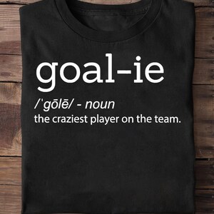 May include: Black t-shirt with white text that reads "goal-ie /'gōlē/ - noun the craziest player on the team."