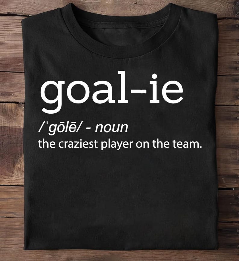 May include: Black t-shirt with white text that reads "goal-ie /'gōlē/ - noun the craziest player on the team."