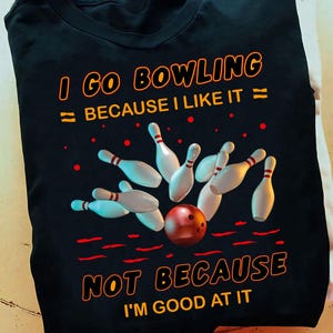 K&ouml;nnte beinhalten: Schwarzes T-Shirt mit einem Bowling-Motiv. Das Motiv zeigt eine Bowlingkugel und Pins mit dem Text "Ich gehe Bowling, weil ich es mag, nicht weil ich gut darin bin."