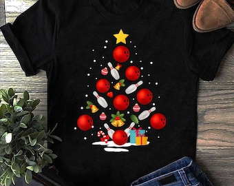 May include: Black t-shirt with a Christmas tree design composed of red bowling balls, white bowling pins, and gold bells. A yellow star tops the tree, with gifts and candy canes at the base. The shirt is displayed on a wooden surface.