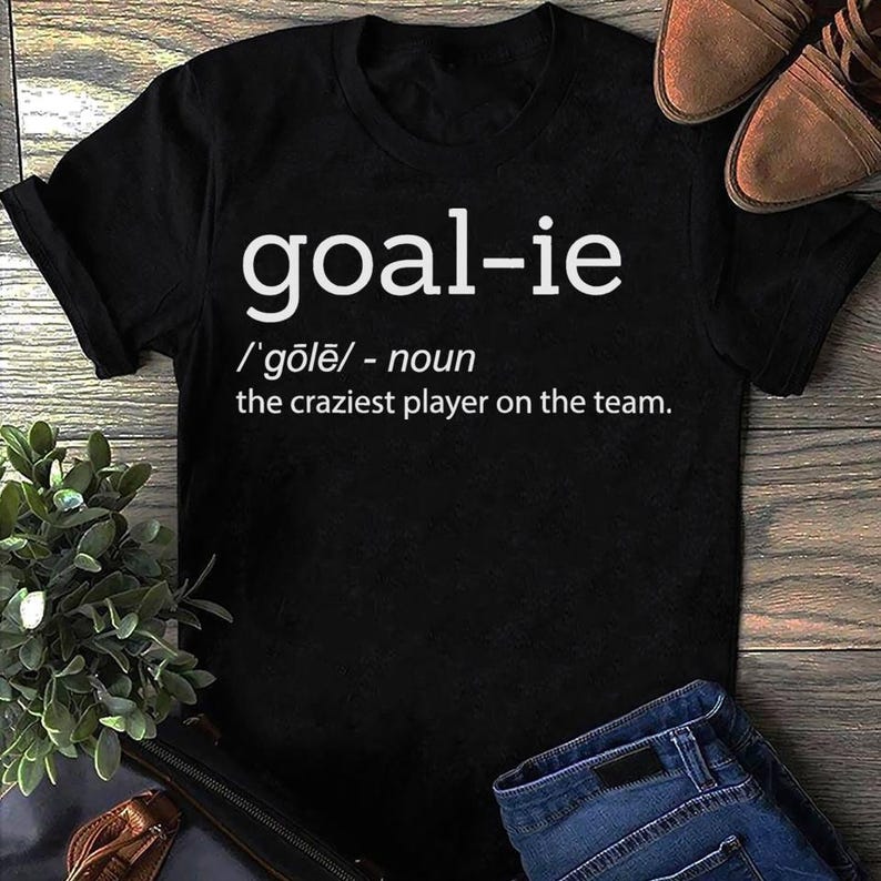 May include: Black t-shirt with white text that reads "goal-ie /'gōlē/ - noun the craziest player on the team."