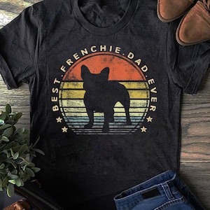 Puede incluir: Una camiseta negra con un gráfico de estilo vintage de una silueta de Bulldog Francés frente a una puesta de sol. El texto "Best Frenchie Dad Ever" está escrito en un círculo alrededor del perro.
