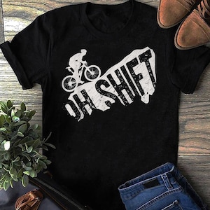 Könnte beinhalten: Schwarzes T-Shirt mit einer weißen Grafik einer Person, die mit dem Mountainbike einen Hügel hinauffährt, mit dem Text "Oh Shift" in einer verwitterten Schrift.