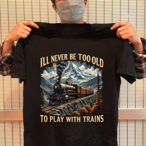 May include: Black t-shirt with a train graphic and the text "I'll Never Be Too Old To Play With Trains." The graphic features a vintage train traveling through a mountain landscape with a snowy peak and trees.