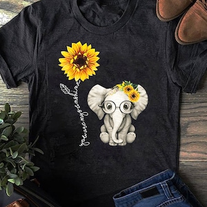 May include: A black t-shirt with a white elephant wearing glasses and a sunflower crown. The elephant is standing next to a sunflower and the text "you are my sunshine" is written vertically beside the elephant.