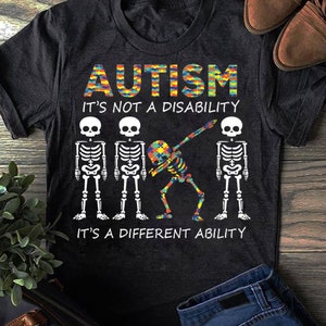 May include: Black t-shirt with a colorful puzzle piece design that says "AUTISM IT'S NOT A DISABILITY IT'S A DIFFERENT ABILITY". Four white skeleton figures are below the text, with the middle skeleton doing a dab.