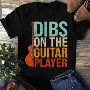 May include: Black t-shirt with a vintage-style graphic of a guitar and the text "DIBS ON THE GUITAR PLAYER".
