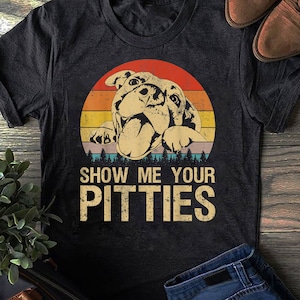 Pode incluir: Uma camiseta preta com um gráfico de pôr do sol de estilo vintage com um pit bull. O texto "Show Me Your Pitties" é impresso abaixo do gráfico.
