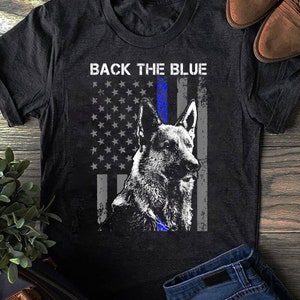 Peut inclure: Un t-shirt noir avec un drapeau américain à ligne bleue et un berger allemand. Le texte "BACK THE BLUE" est imprimé sur le t-shirt.