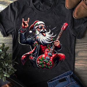 Puede incluir: Camiseta gris oscuro con un gráfico de Papá Noel con chaqueta de cuero negra, gafas de sol y gorro de Papá Noel, tocando una guitarra roja. Papá Noel hace el gesto de la mano de rock. La guitarra está decorada con adornos navideños.