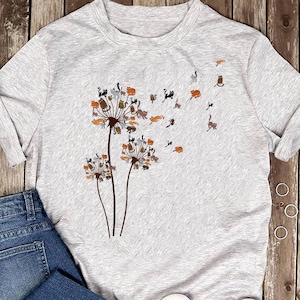 Könnte beinhalten: Hellgraues T-Shirt mit einem Löwenzahnmotiv und Katzensilhouetten. Die Löwenzahnhäupter sind braun und beige, mit orangefarbenen, schwarzen und braunen Katzenformen. Die Katzen scheinen im Wind zu wehen. Das T-Shirt hat einen Rundhalsausschnitt und kurze Ärmel.