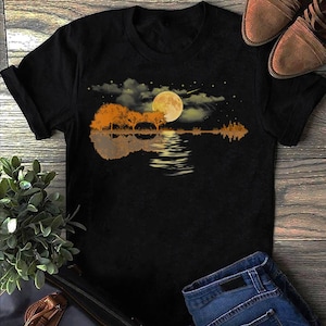 Può includere: Maglietta nera con una grafica di una silhouette di chitarra riflessa in un lago con luna piena e stelle nel cielo.