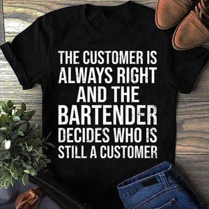 Op de afbeelding: Zwart t-shirt met witte tekst: "THE CUSTOMER IS ALWAYS RIGHT AND THE BARTENDER DECIDES WHO IS STILL A CUSTOMER." De afbeelding toont ook bruine schoenen en een blauwe jeans.