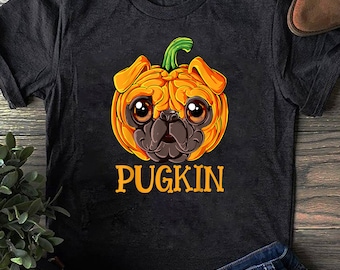 Puede incluir: Una camiseta negra con un pug de dibujos animados que lleva un disfraz de calabaza. El texto "PUGKIN" está impreso debajo de la imagen.