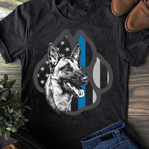 May include: A black t-shirt featuring a graphic of a German Shepherd dog's head in front of an American flag with a blue line running down the centre. The graphic is in the shape of a paw print.