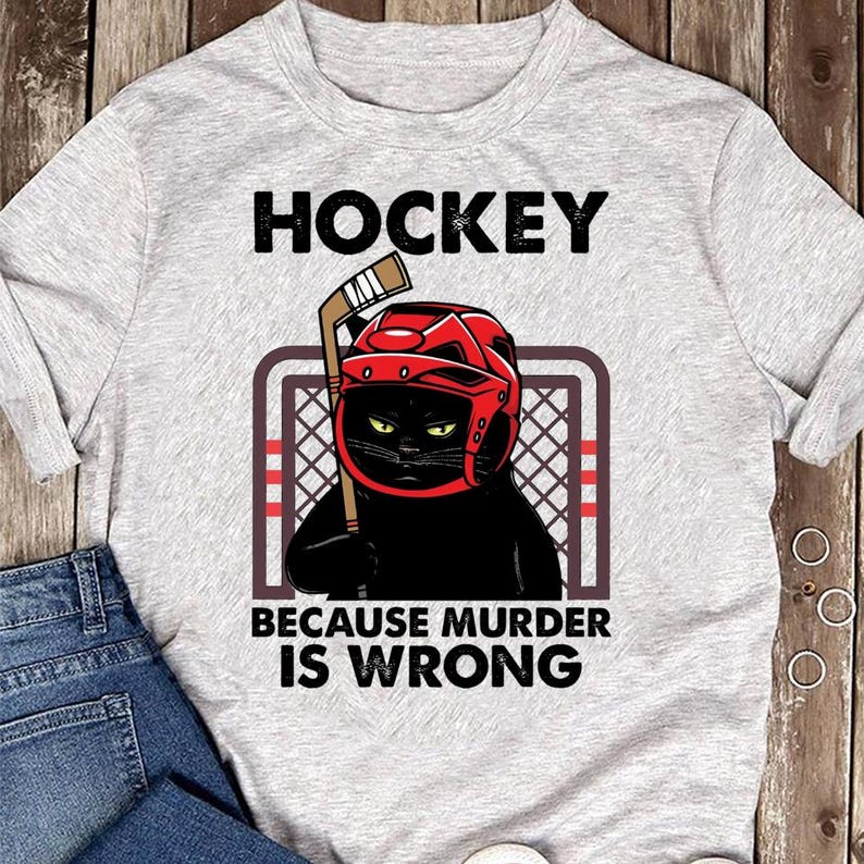 May include: Light gray t-shirt with the word "HOCKEY" in black block letters. Below is a graphic of a black cat wearing a red hockey helmet and holding a hockey stick. The text "BECAUSE MURDER IS WRONG" is below the cat.