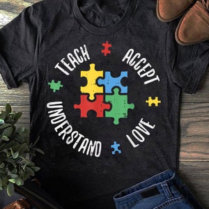 Könnte beinhalten: Ein schwarzes T-Shirt mit einem weißen Grafikdesign, das ein Puzzle-Teil mit den Worten "Teach, Accept, Understand, Love" zeigt.
