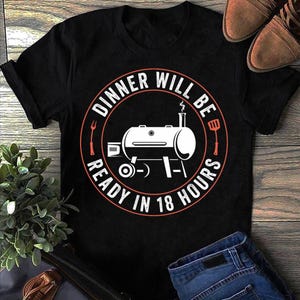May include: Black t-shirt featuring a circular design with the text "Dinner Will Be Ready In 18 Hours." The graphic includes a white barbecue grill illustration with red details. The image also shows brown shoes and blue jeans.