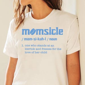 Puede incluir: Camiseta blanca con la palabra "momsicle" en azul claro y una definición. La definición dice: "alguien que se para en una pista de hielo y se congela por el amor de su hijo."