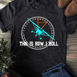 Może przedstawiać: Czarny T-shirt z białym kompasem i niebieskim nadrukiem samolotu. Kompas wskazuje kurs 45 stopni. Pod kompasem znajduje się napis "THIS IS HOW I ROLL".