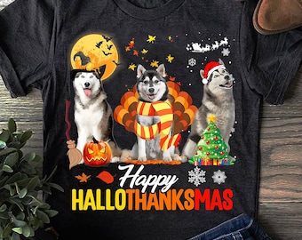 Puede incluir: Camiseta gris oscuro con un diseño festivo que presenta a tres Huskies siberianos. Los perros están vestidos para Halloween y Navidad. El texto "Happy HALLOTHANKSMAS" se muestra debajo.