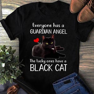 Może przedstawiać: Czarny t-shirt z napisem "Everyone has a GUARDIAN ANGEL. The lucky ones have a BLACK CAT." Czarny kot z żółtymi oczami i czerwonym sercem jest na koszulce.