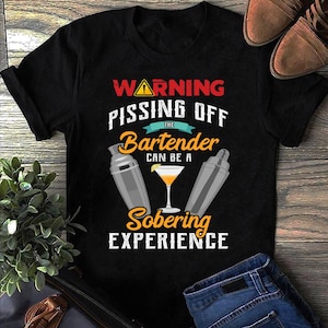 Op de afbeelding: Zwart T-shirt met de tekst "WARNING PISSING OFF THE Bartender CAN BE A Sobering EXPERIENCE". Het ontwerp bevat cocktailshakers en een cocktailglas met een schijfje citroen. Het shirt wordt gecombineerd met een blauwe jeans en bruine schoenen.
