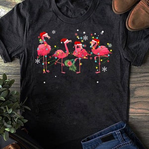 May include: A black t-shirt with a Christmas-themed design featuring four pink flamingos wearing Santa hats and decorated with colorful Christmas lights.
