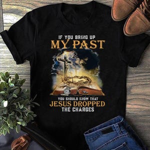 Puede incluir: Camiseta negra con un gráfico de una cruz y un libro abierto. El texto en la camiseta dice "Si sacas a relucir mi pasado, debes saber que Jesús dejó caer los cargos."