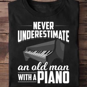 May include: Black t-shirt featuring the text "NEVER UNDERESTIMATE an old man WITH A PIANO" in white. A grayscale illustration of a piano is below the text. The shirt is folded on a wooden surface.