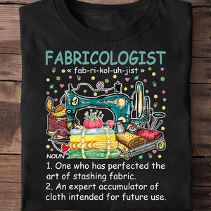 May include: Black t-shirt featuring a sewing machine and fabric illustration. The text reads "FABRICOLOGIST" with a definition below, including the phrase "art of stashing fabric".