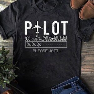 May include: Black t-shirt with white text that reads "Pilot in Progress" with a graphic of a plane and a progress bar with three planes. The text "Please Wait..." is below the progress bar.
