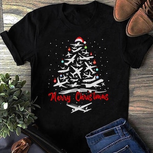 May include: Black t-shirt featuring a white Christmas tree design made of aeroplanes. The tree is decorated with colourful baubles and a red Santa hat. The text "Merry Christmas" is printed below the tree.