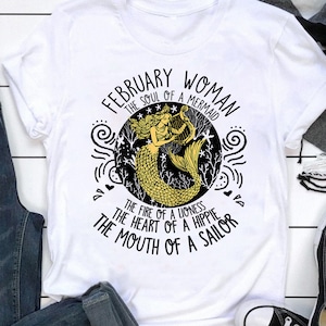 May include: White t-shirt with a black and gold graphic design featuring a mermaid playing a harp. The text reads "February Woman The Soul of a Mermaid The Fire of a Lioness The Heart of a Hippie The Mouth of a Sailor"
