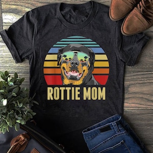 Puede incluir: Una camiseta negra con un gráfico de estilo vintage de un Rottweiler con gafas de sol frente a una puesta de sol. El texto "Rottweiler Mom" está impreso debajo del gráfico.