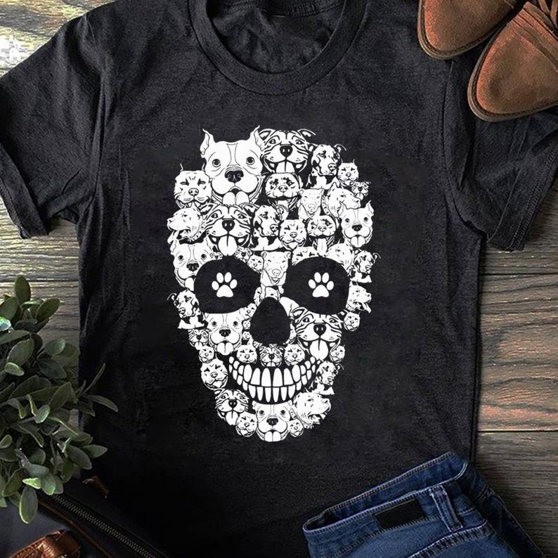May include: A black short-sleeved t-shirt featuring a large white graphic print on the front. The design is a skull shape composed of numerous small illustrations of dog faces, with two white paw prints forming the eye sockets of the skull.