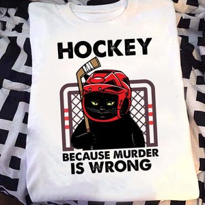 May include: White t-shirt with a black cat wearing a red hockey helmet and holding a hockey stick. The shirt has the word "HOCKEY" above the cat and "BECAUSE MURDER IS WRONG" below.