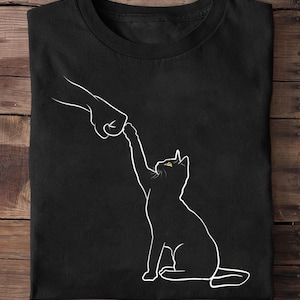 K&ouml;nnte beinhalten: Schwarzes T-Shirt mit einer wei&szlig;en Umrissillustration einer Katze, die nach einer Hand greift. Die Katze hat gelbe Augen. Das Design ist auf dem Shirt zentriert.