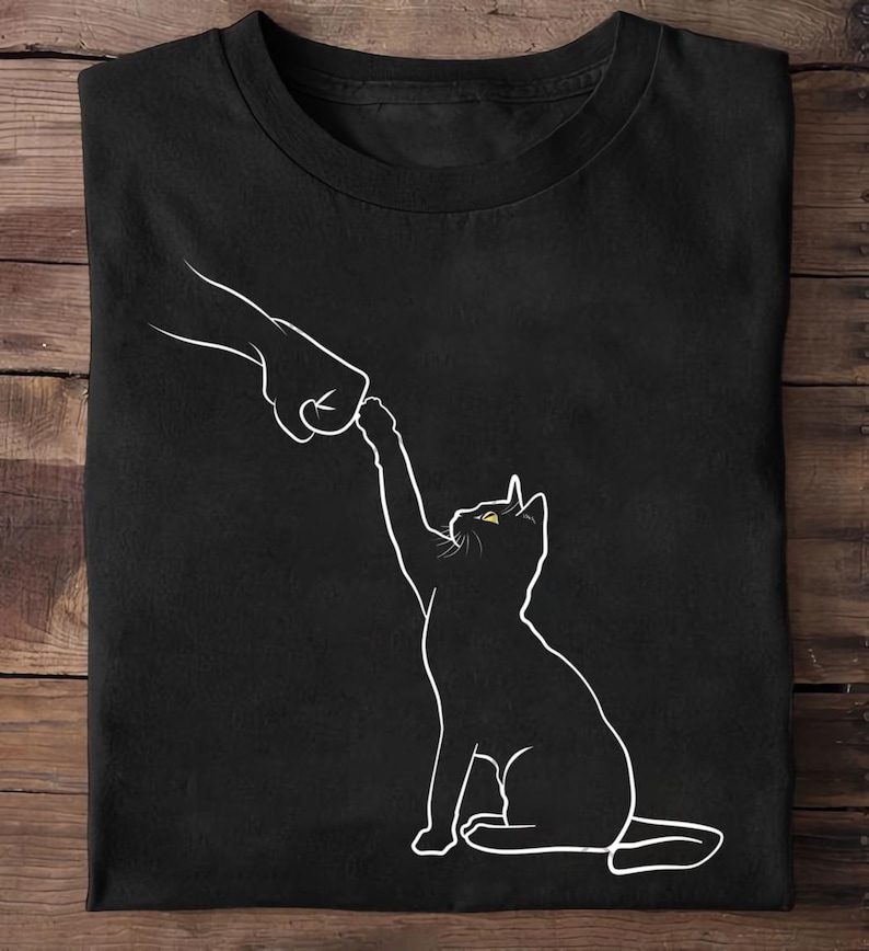 K&ouml;nnte beinhalten: Schwarzes T-Shirt mit einer wei&szlig;en Umrissillustration einer Katze, die nach einer Hand greift. Die Katze hat gelbe Augen. Das Design ist auf dem Shirt zentriert.