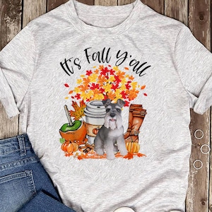 Puede incluir: Una camiseta gris con el texto "It's Fall y'all" impreso en ella. El diseño presenta un pequeño perro gris, una taza de café, hojas de otoño y botas.