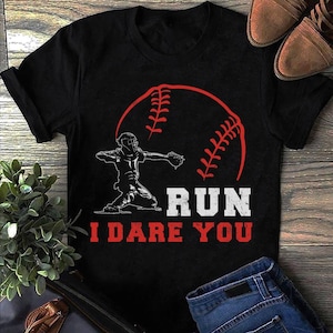 Puede incluir: Camiseta negra con un jugador de béisbol blanco atrapando una pelota de béisbol roja. El texto "RUN I DARE YOU" está impreso en blanco en la camiseta.